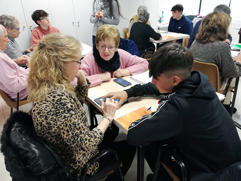 Un jove ensenya el funcionament d'un telèfon a dues sòcies de la llar de la Gent Gran. (Foto: Ajuntament de Palamós).